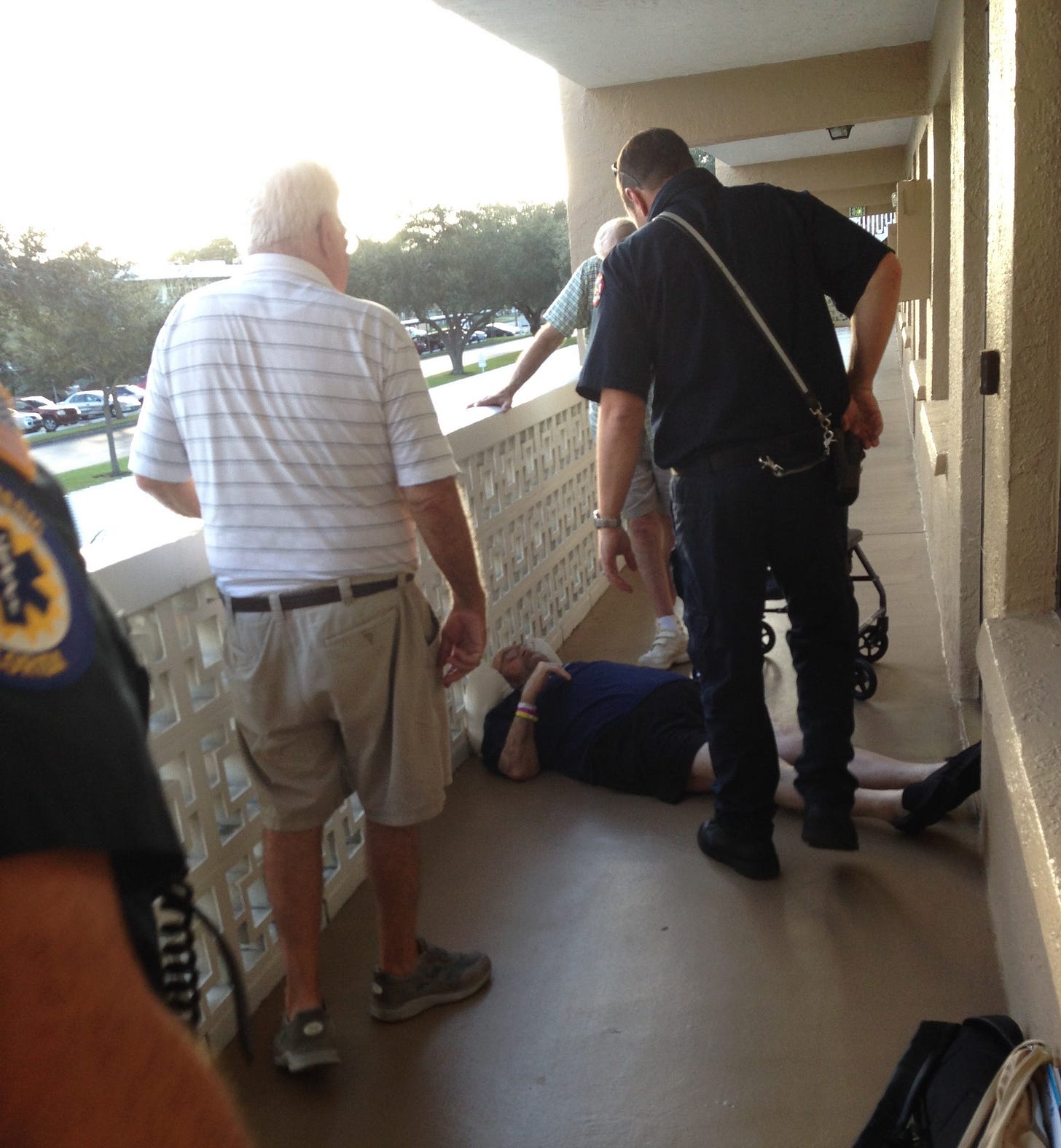 An elderly man lays on the ground in a hallway surrounded by an EMT and other onlookers.