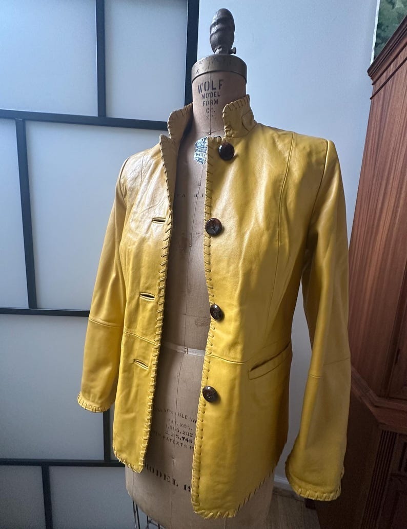 May include: A yellow leather jacket with a mandarin collar and button closure. The jacket has stitching detail around the edges and pockets. The jacket is on a mannequin.