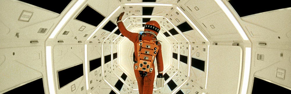 Production still from 1968 sci-fi film 2001. An astronaut in an orange space suit walks away from the camera along a long, symmetrical, tunnel-like corridor on a space ship