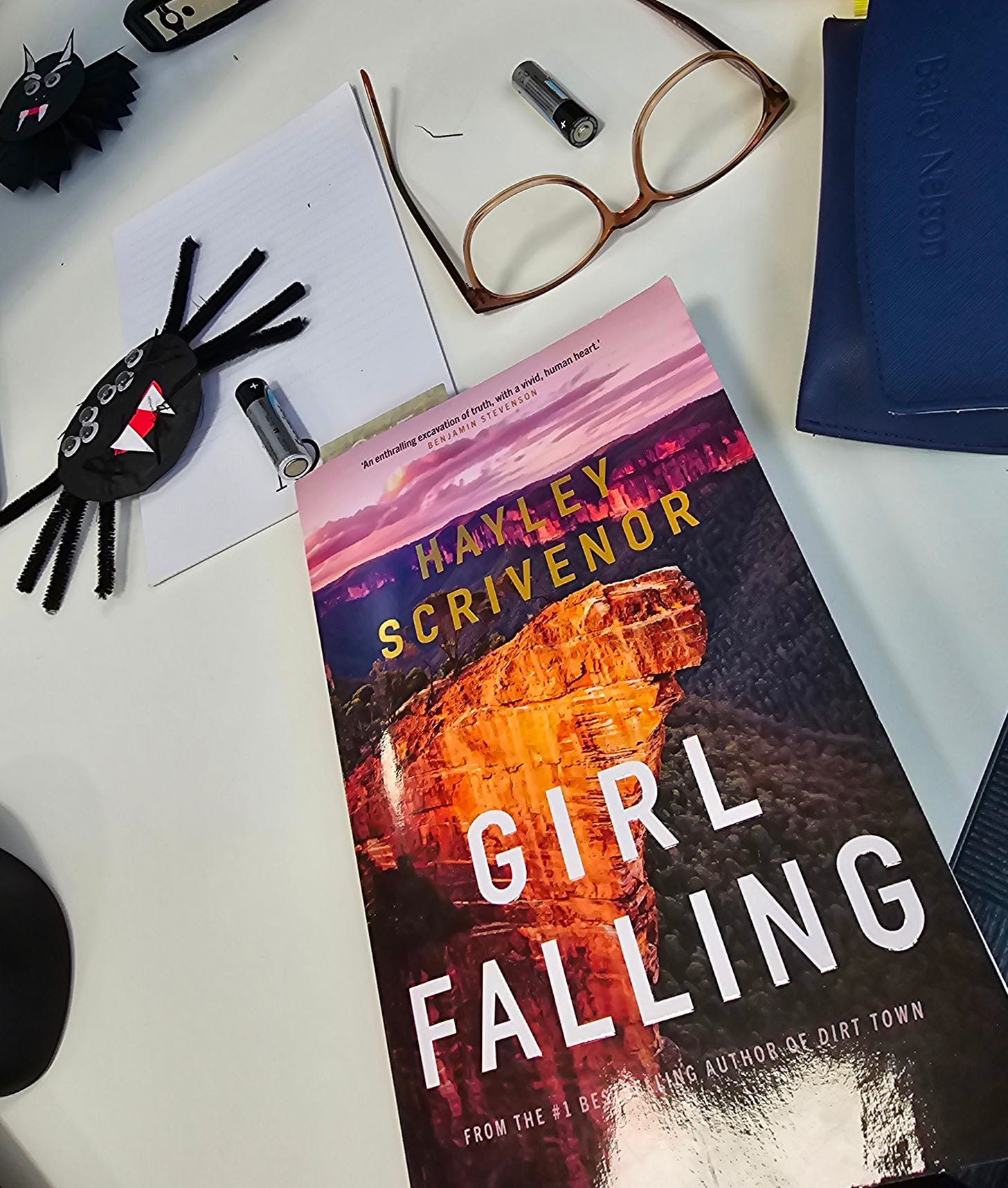 It's a work desk. On it there's a pair of glasses, a couple of very janky looking homemade Halloween decorations, and a copy of the book Girl Falling by Hayley Scrivenor.