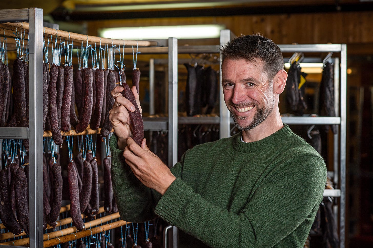 Adrian Hirt steht im Reiferaum der Fleischtrocknerei Sialm und hält eine getrocknete Wurst hoch, die an einer blauen Schnur hängt. Um ihn herum hängen auf mehreren Holzstangen Dutzende weiterer dunkelrot gereifter Würste. Der Hintergrund zeigt Metallgestelle und warmes Werkstattlicht.