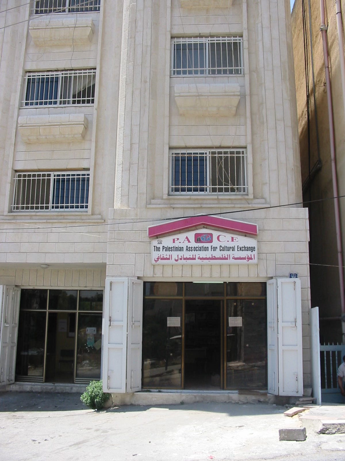 Exterior of a multi-story limestone building in al-Bireh, West Bank, with a bilingual sign reading "PACE — The Palestinian Association for Cultural Exchange" in English and Arabic. The entrance doors are open. Photographed in June 2004.