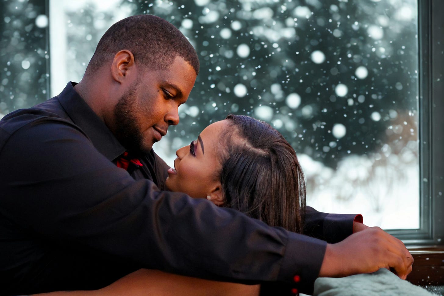 A Black couple sits close together by a window as snow falls outside. The man leans in with a gentle, loving expression, while the woman looks up at him with a soft smile. Their foreheads nearly touch, creating an intimate, tender moment framed by the winter scene behind them.