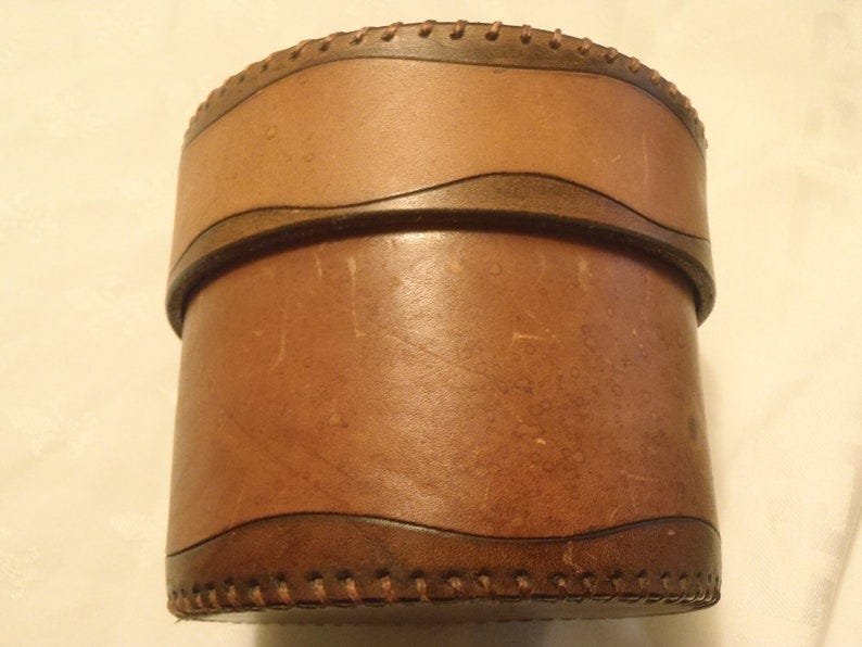 May include: A brown leather cylindrical box with a stitched edge. The box has a brown leather band around the top and bottom with a wavy design.