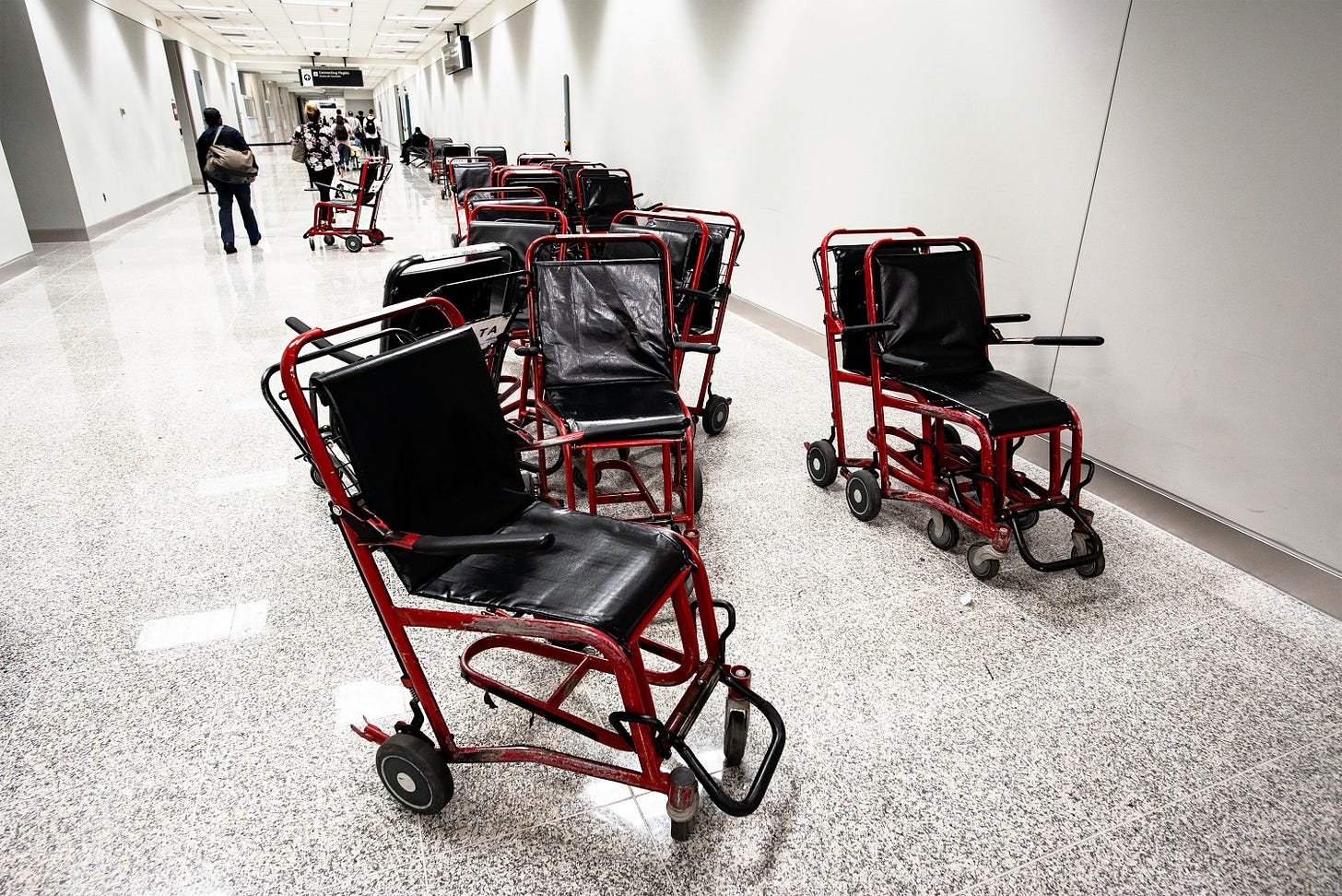 A row of red-framed airport transfer wheelchairs with black vinyl seats, parked along a wide airport corridor. 