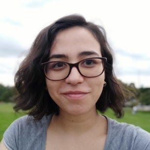 #PraTodosVerem: Fernanda é uma mulher branca de cabelos escuros na altura dos ombros, levemente ondulados. Ela usa óculos de grau de armação escura e uma camiseta cinza. Ao fundo, um gramado verde, árvores e o céu com nuvens. Fim da descrição. 