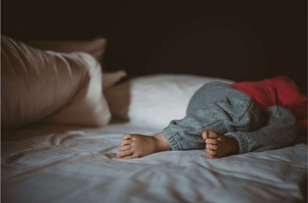 Photo from Canva by jenellebotts. It's a photo inside a dimly lit bedroom of a child sleeping on a bed. The pillows are sideways and the duvet pulled back.