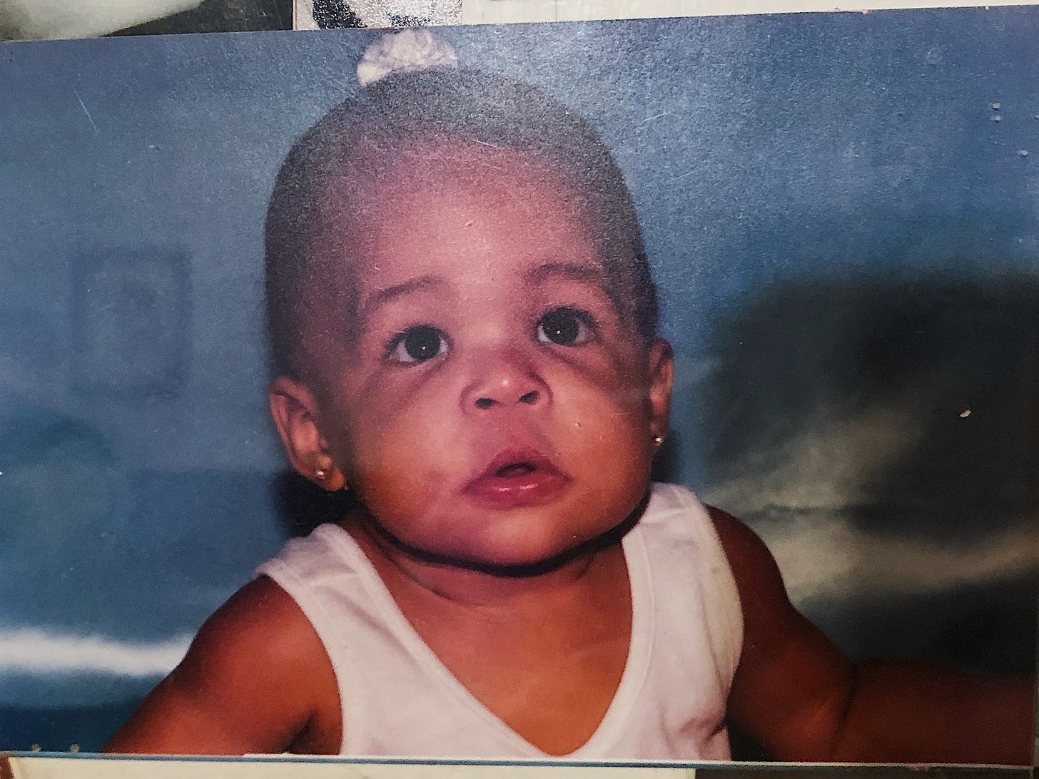 A physical photo of a baby in a white V neck and a little white scrunchy, looking up and over the camera.
