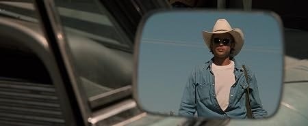 Homem com chapéu de cowboy e óculos escuros refletido no espelho de um carro.