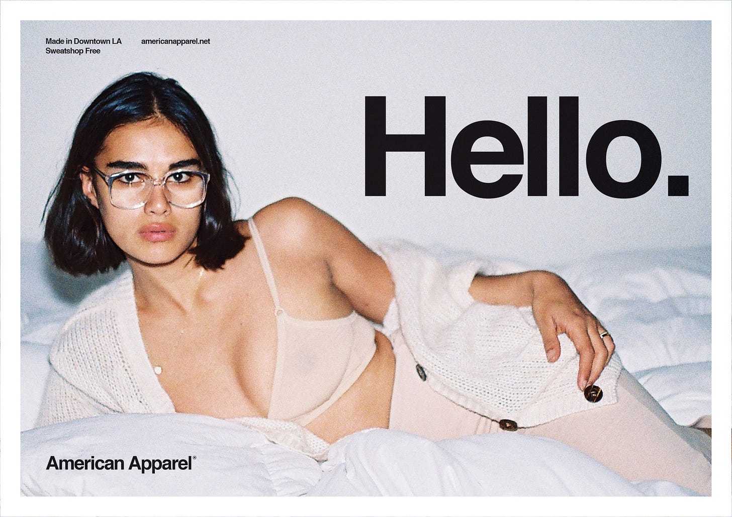 American Apparel's pornified boyfriend cardigan ad, with a model splayed across a bed, cardigan open, bra showing