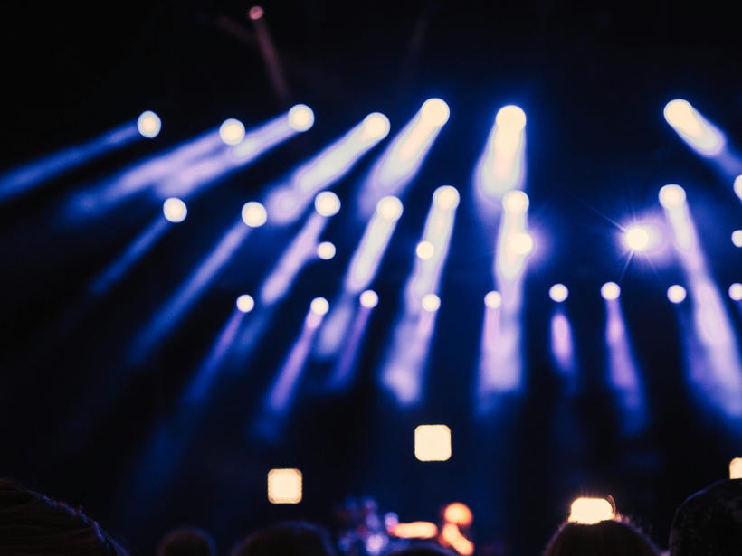 Against a dark background, bright stage lights shine toward the viewer, as though we're at a concert. The picture is mostly blurry and impressionistic, but near the bottom of the frame it looks like people are holding up their phones to record.