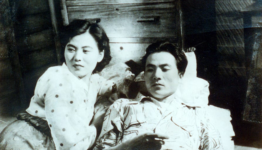 A still from the 1955 film The Widow by Nam-Ok Park. A mother in her late 30s wearing a hanbok is looking off to the side while lounging next to a casually dressed man, who is laying down.