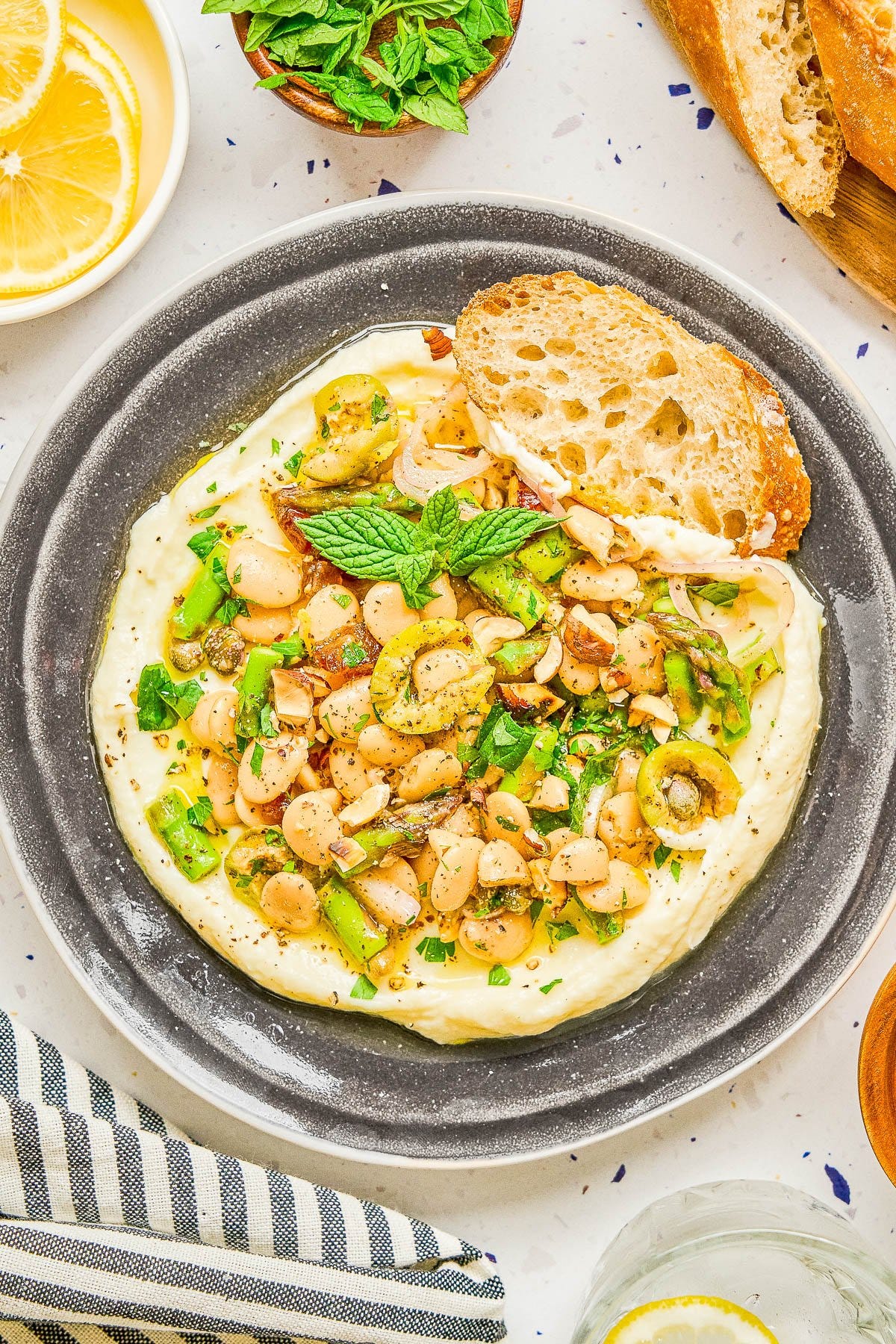 A plate of creamy white hummus topped with chopped green olives, white beans, herbs, and spices, served with a slice of crusty bread.