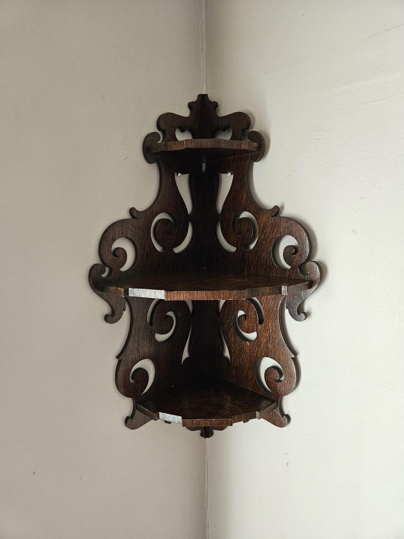May include: A dark brown wooden corner shelf with intricate scrollwork and two shelves. The shelf is mounted on a white wall.
