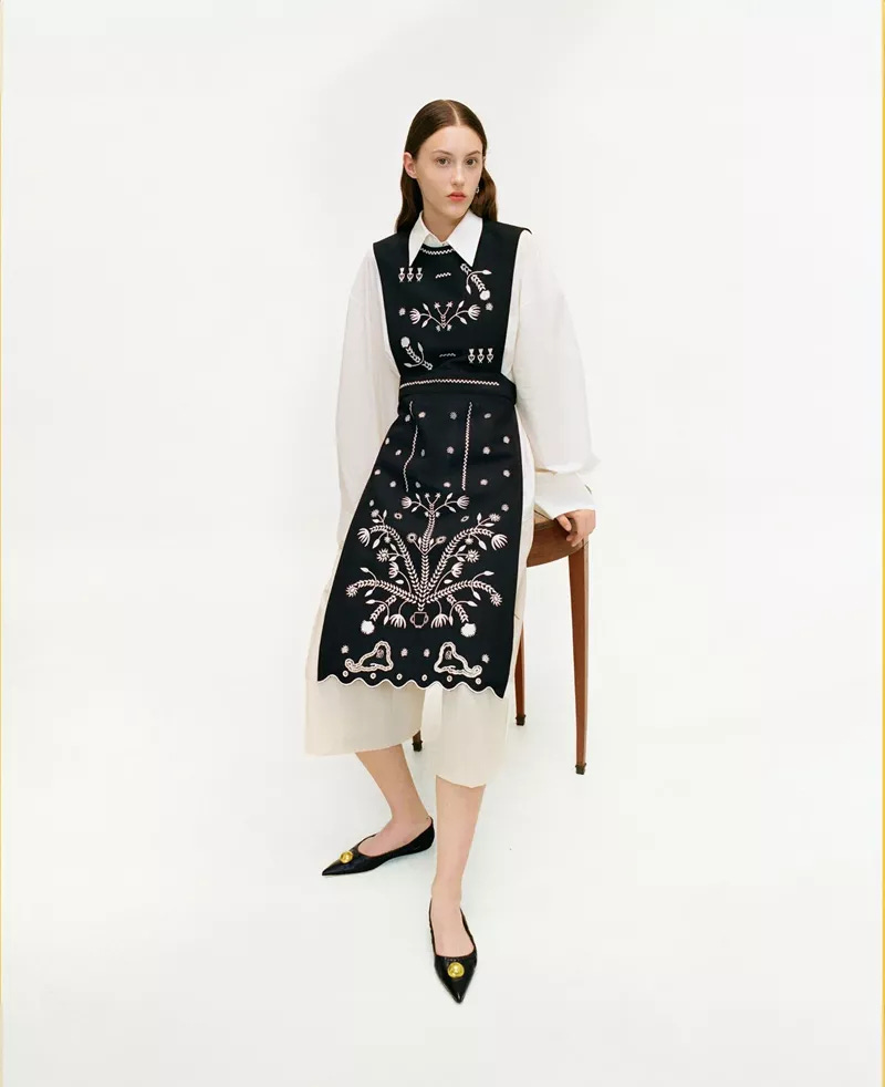 a model wears a white outfit with a black embroidered apron. she leans partially back against a stool
