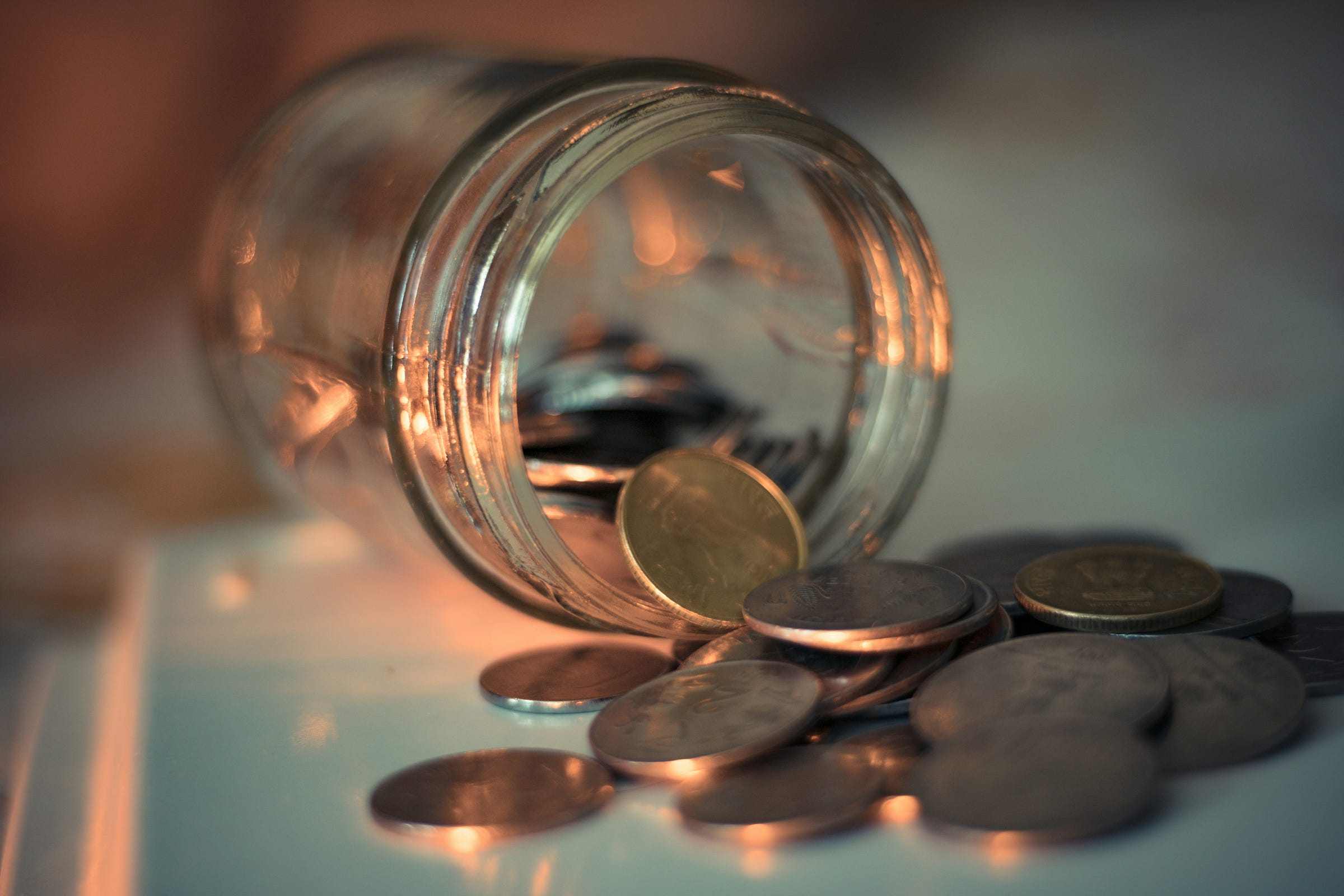The photo of a glass jar tipped over with coins spilling out onto a white surface, lit by warm ambient light The photo of a glass jar tipped over with coins spilling out onto a white surface, lit by warm ambient light