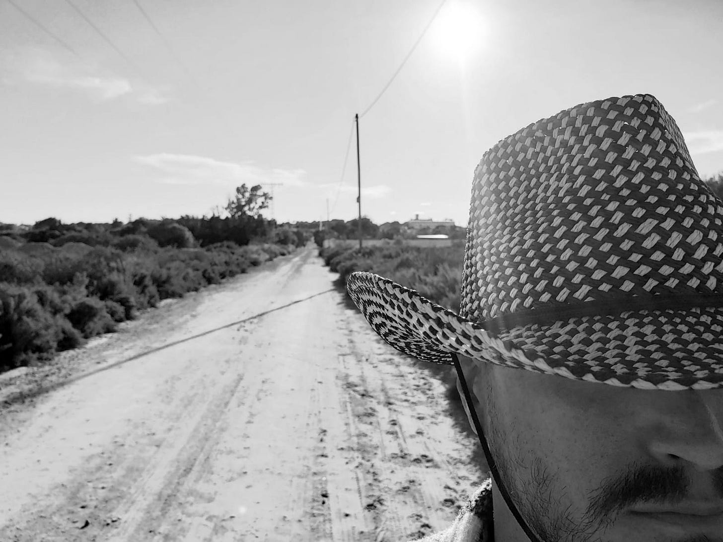 A black and white photo with a man wearing a cowboy hat on the right with a dusty road opening up behind him on a low-shrub landscape.