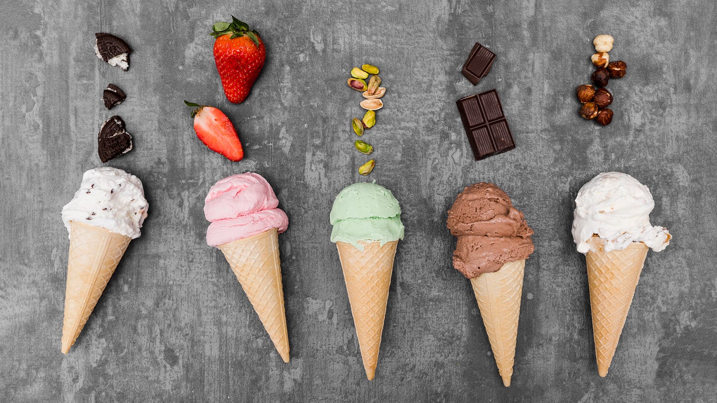 Five ice cream cones on a gray surface. Left to right: white ice cream sprinkled with chocolate and topped with crushed oreo; pink ice cream topped with strawberries; green strawberry with pistachos; brown ice cream topped with pieces of a chocolate bar, and cream ice cream topped with hazelnuts. 