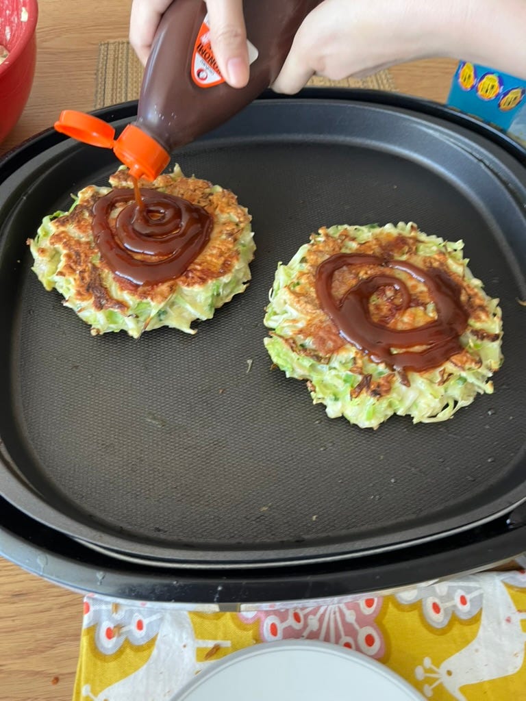 2 browned pancakes with green vegetables inside on a tabletop griddle. There is a set of hands pouring brown sauce out of a container onto one of the pancakes