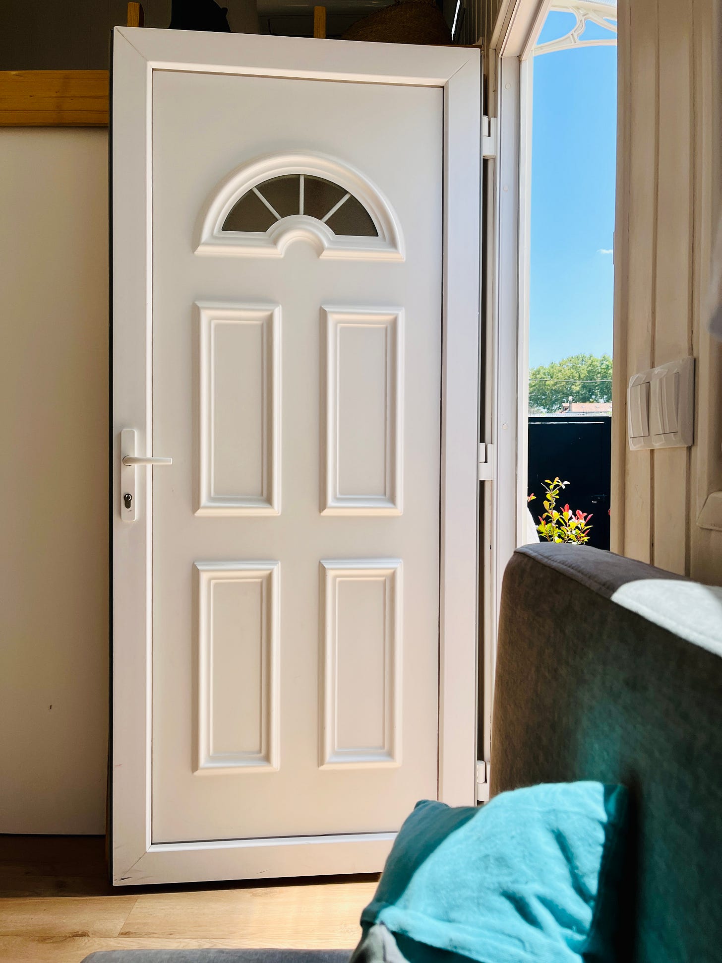 Image: Photo of tiny house main door entrance. A white door, opened, welcoming the sunshine in, shining on the cosy, wooden floor. In the foreground, a glimpse of the grey sofa, with pops of turquoise from the little throw cushions.