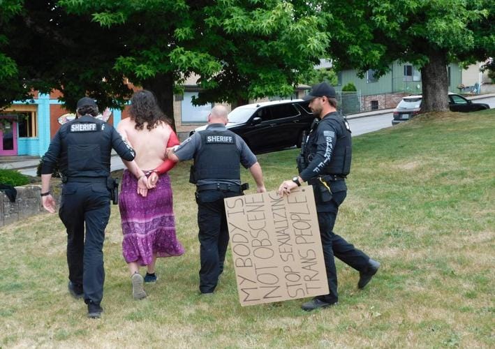 Lucy Lauser, topless and handcuffed with one arm painted red, is escorted across a grass lawn by three Skamania County Sheriff's deputies. A deputy carries her cardboard protest sign, which reads "MY BODY IS NOT OBSCENE / STOP SEXUALIZING TRANS PEOPLE."