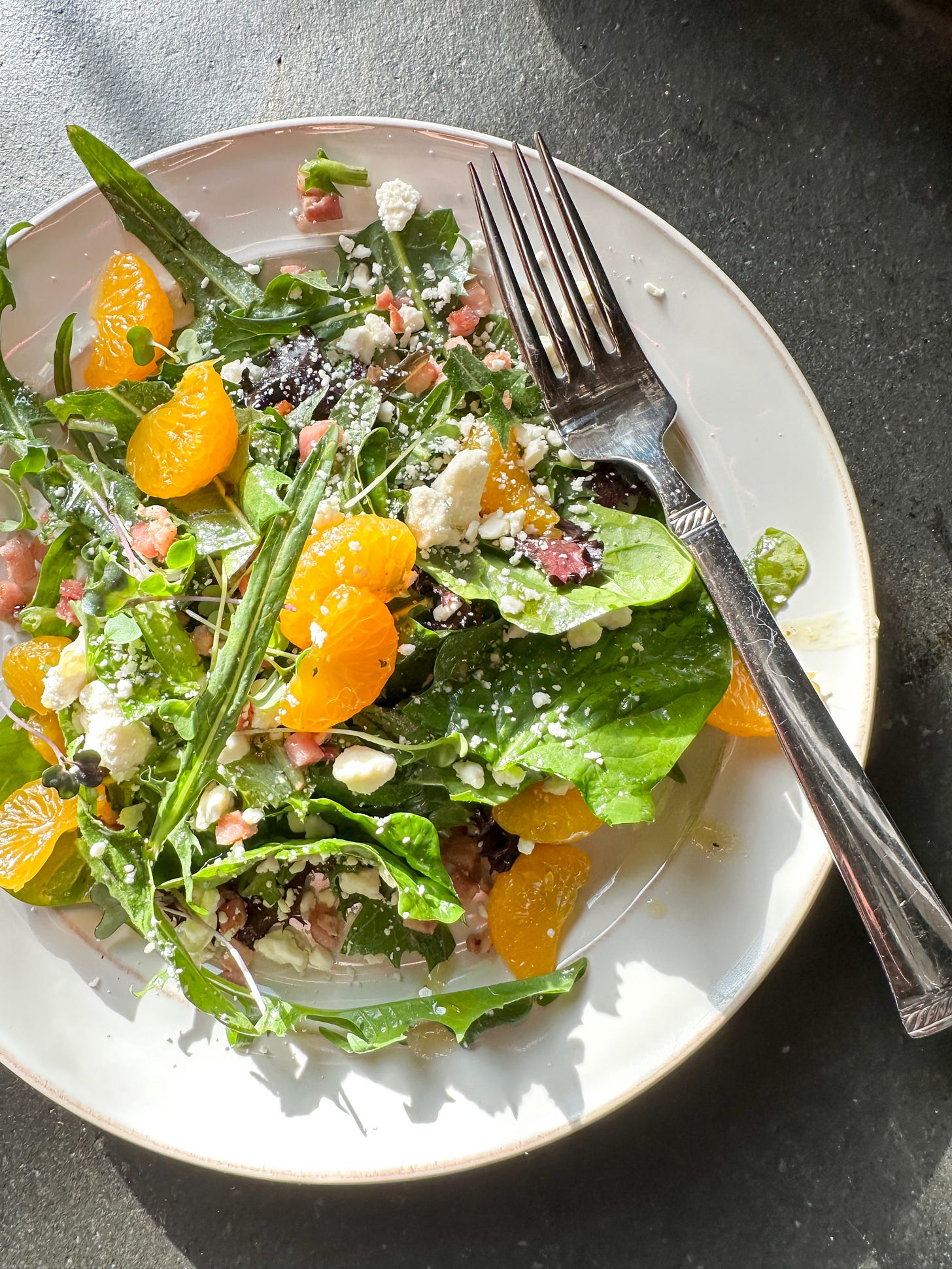 White plate with spring greens, mandarin oranges, crumbled goat cheese, crispy pancetta in a honey vinaigrette