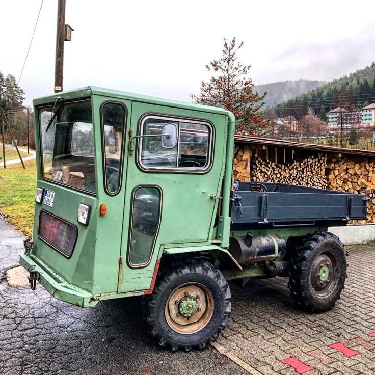 This may contain: an old green truck is parked on the side of the road in front of a pile of logs