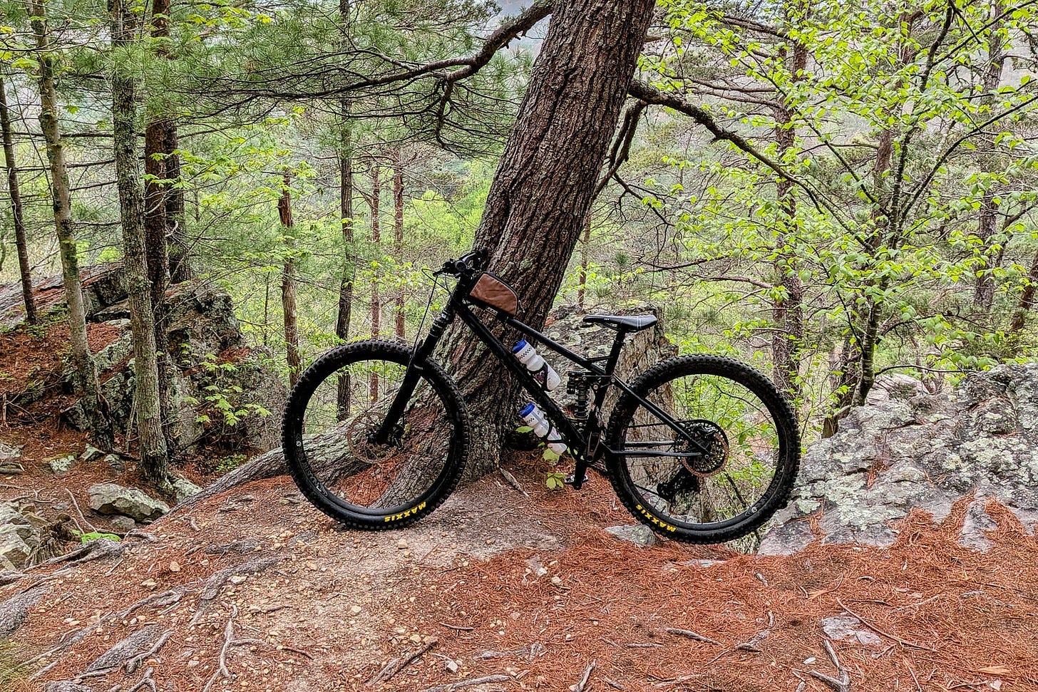 A full-suspension mountain bike leans against a tree on a rocky forest trail, surrounded by pine needles, stones, and dense green foliage. A full-suspension mountain bike leans against a tree on a rocky forest trail, surrounded by pine needles, stones, and dense green foliage.