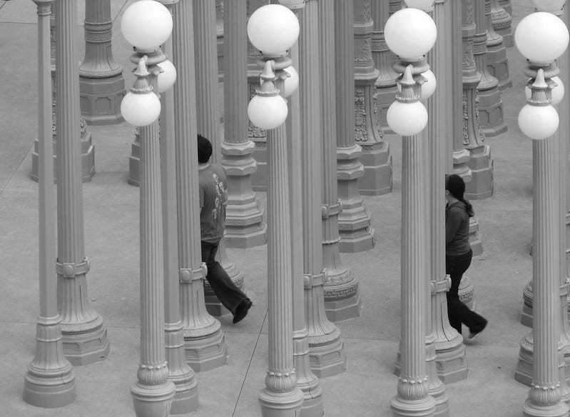 Monochrome photo looking down on Chris Burden's Urban Light installation at the Los Angeles County Museum of Art on Wilshire Boulevard in Los Angeles, consisting of more than 200 restored street lamps from the 1920s and 1930s. The lamp poles are arranged in a grid outside the museum entrance. Two museum goers are walking through the installation in step but separated and partially obscured.