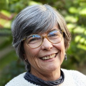 Fotografia em close mostra a mulher sorrindo abertamente. Ela tem cabelos grisalhos curtos e óculos de armação âmbar, e usa o suéter de tricô texturizado com a gola rulê cinza. O fundo verde e borrado sugere um ambiente de folhas e flores.