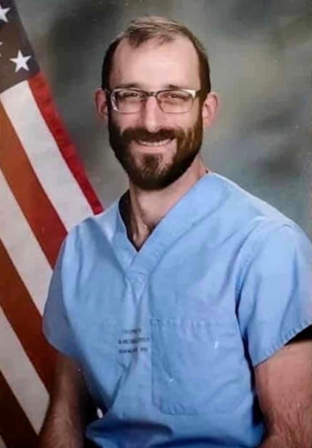 Alex Pretti -- a smiling man in glasses and blue scrubs and a well-trimmed brown beard -- smiles in an official portrait. He stands in front of an American flag.