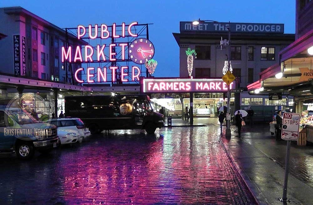 This is a photograph of Pike Place Market in Seattle at dusk or dawn. The neon signs for "PUBLIC MARKET CENTER" and "FARMERS MARKET" are brightly lit, casting a vivid pink and purple reflection on the wet cobblestone street. The street appears to be wet from rain. In the background, a dark delivery truck and several cars are parked, and people with umbrellas can be seen walking. A sign on a building reads "MEET THE PRODUCER," and another, to the left, says "LA SALLE HOTEL."