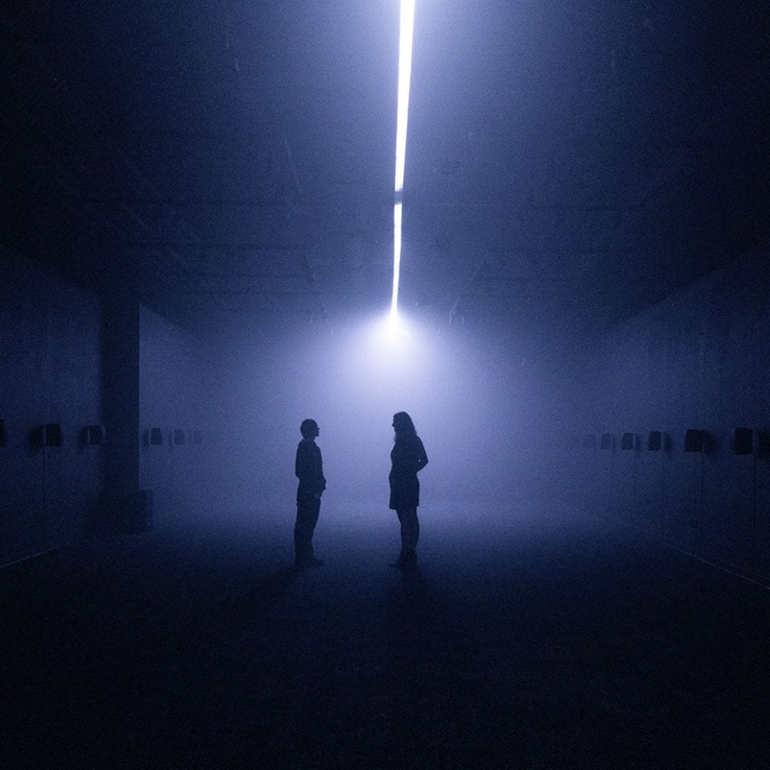 A photo of a dark room with a light down the center. You can see speakers along both walls and two people standing in the center of the room backlit and in shadow. A photo of a dark room with a light down the center. You can see speakers along both walls and two people standing in the center of the room backlit and in shadow.