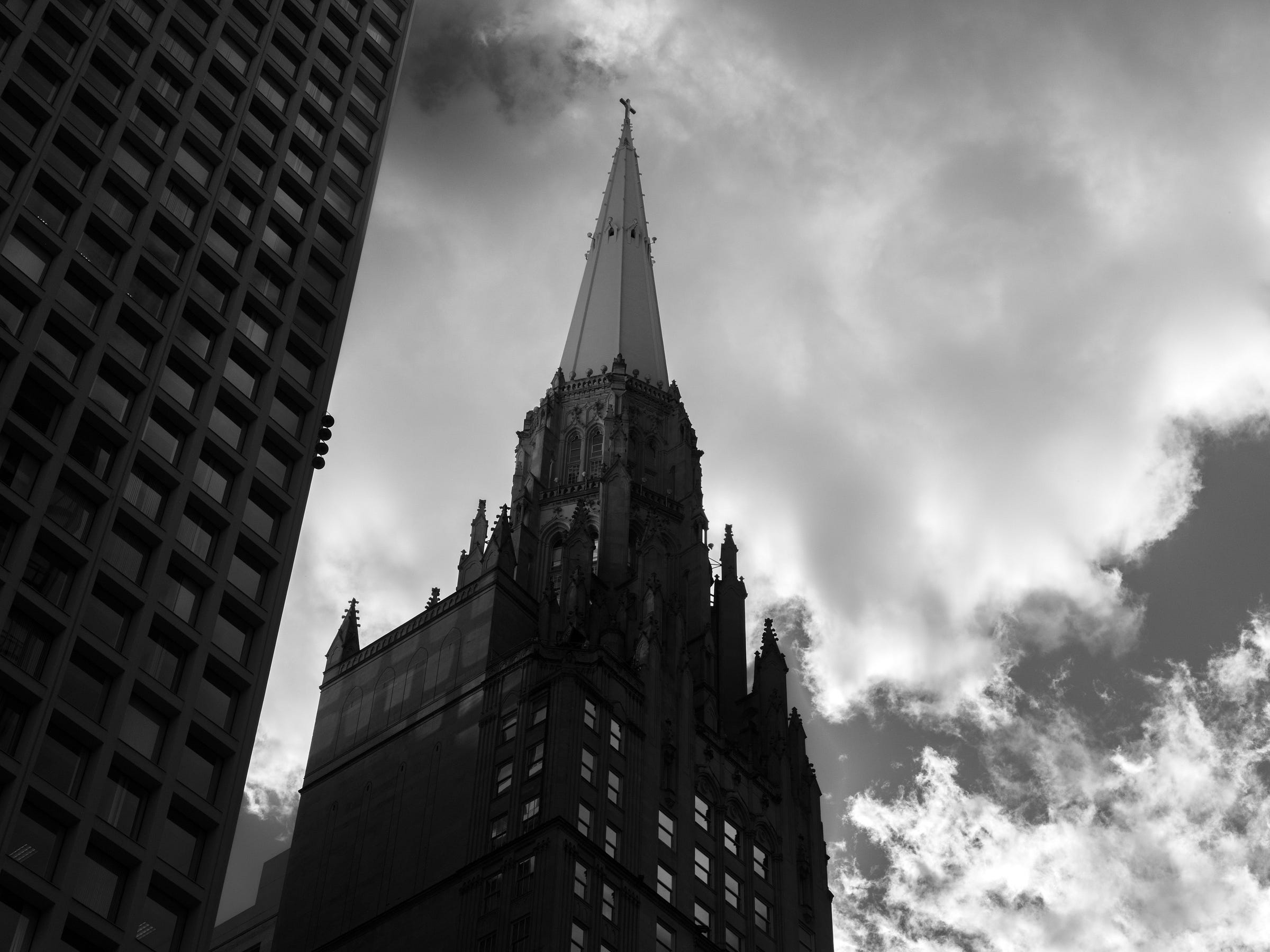 lack and white photograph of a towering neo-Gothic skyscraper with an ornate spire, framed by a modern high-rise on the left. Dramatic clouds swirl in the sky behind, creating a moody contrast between architecture and atmosphere. lack and white photograph of a towering neo-Gothic skyscraper with an ornate spire, framed by a modern high-rise on the left. Dramatic clouds swirl in the sky behind, creating a moody contrast between architecture and atmosphere.