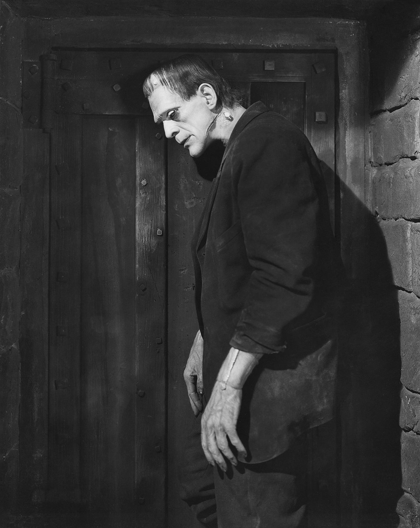 Black-and-white publicity still showing a tall, imposing man with a flat-topped head, heavy facial makeup and neck bolts, standing hunched beside a large wooden door in a stone-walled room. Black-and-white publicity still showing a tall, imposing man with a flat-topped head, heavy facial makeup and neck bolts, standing hunched beside a large wooden door in a stone-walled room.
