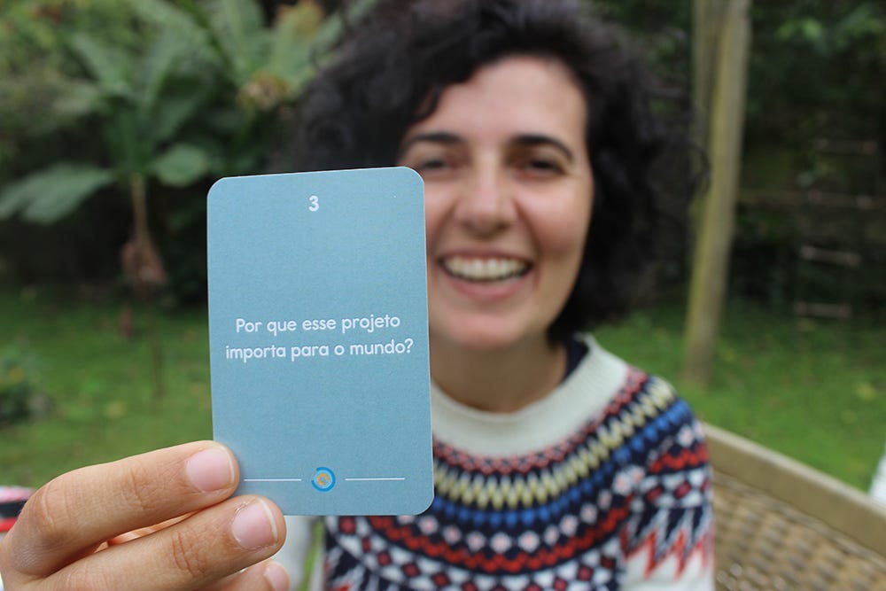 nessa foto a Mari aparece segurando uma carta do Combinados onde se lê "Por que esse projeto importa para o mundo?". Mari é uma mulher branca, de cabelos pretos e enrolados, e está vestindo uma blusa de lã branca com estampas azuis, amarelas e laranjas na gola. ela está sentada em uma cadeira de palha, num jardim com algumas árvores ao fundo.
