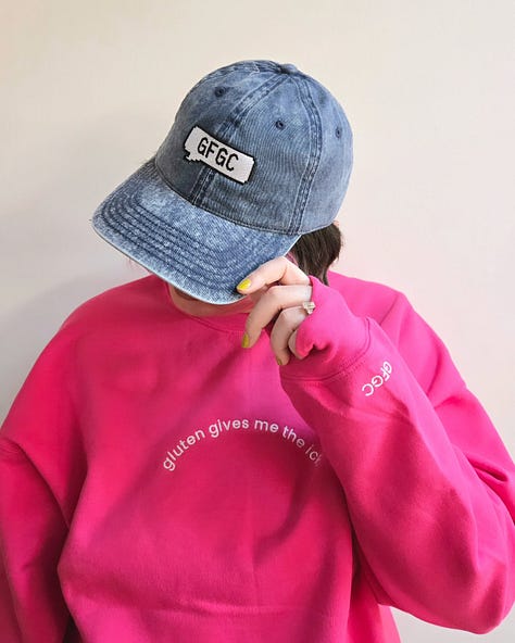 Modeling merch: A hot pink sweatshirt with white embroidered text on the front chest that says "gluten gives me the ick" and "GFGC" on the left sleeve near the wrist. A vintage wash denim baseball cap with an embroidered white pixelated chat bubble and black "GFGC" inside. A black sweatshirt with a green embroidered design on the front chest that says "GAME OVER, YOU ATE GLUTEN AND DIED" with a pixelated ox and wagon and "GFGC" on the left sleeve near the wrist.