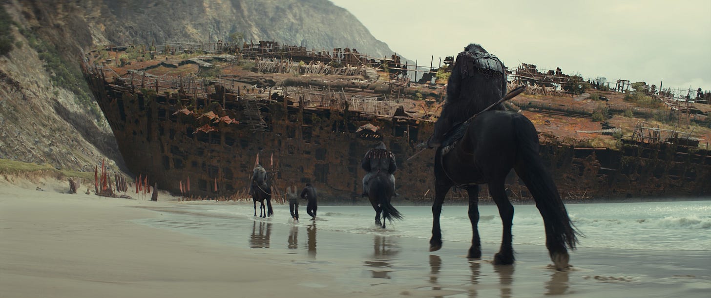 Three apes on horseback and an ape and a human on foot walk in a line on a beach away from the camera, just above the waterline. In the background looms a rusted-out container ship that appears to have been transformed into a dwelling of some sort.