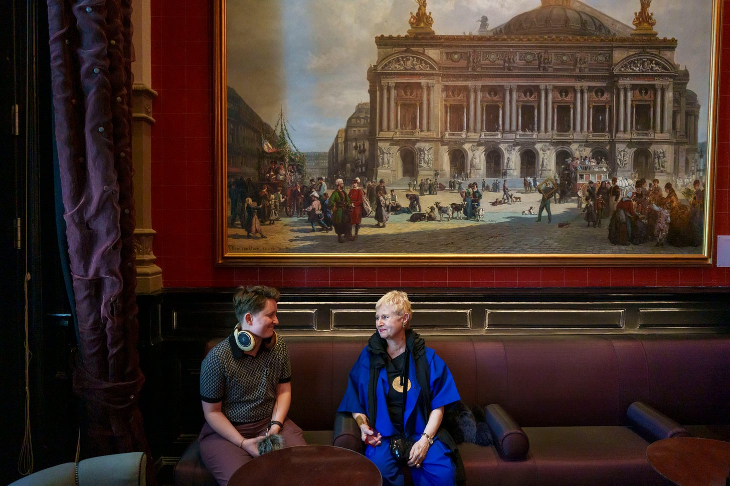 Hannah, with audio equipment, and Marina Mahler, a stylishly dressed octogenarian with short hair, smile at each other. They are sitting in a handsomely appointed room at the Concertgebouw, with a painting of the building itself behind them.