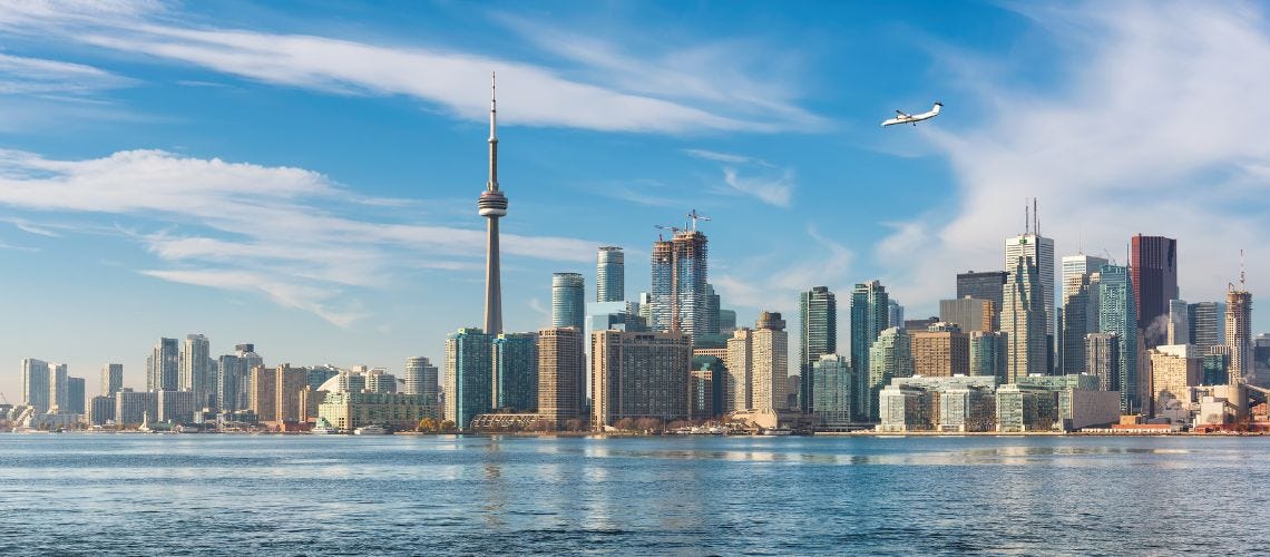 Toronto skyline with the CN Tower, high-rise buildings, and a small plane flying over Lake Ontario under a blue sky.