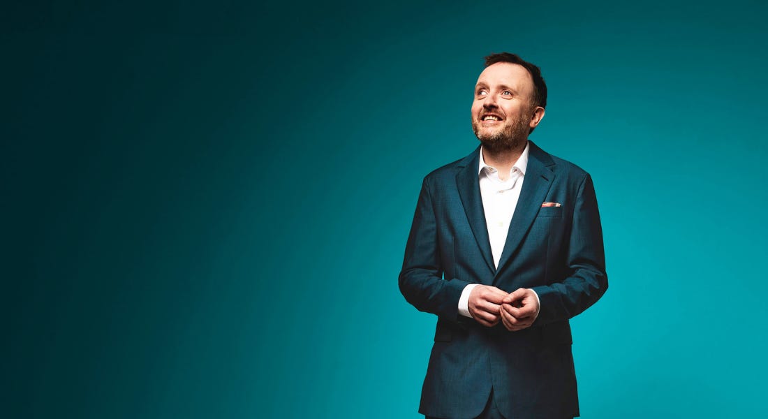 A man - Chris McCausland - wearing a navy blue suit looking up to the left, with a blue background behind him