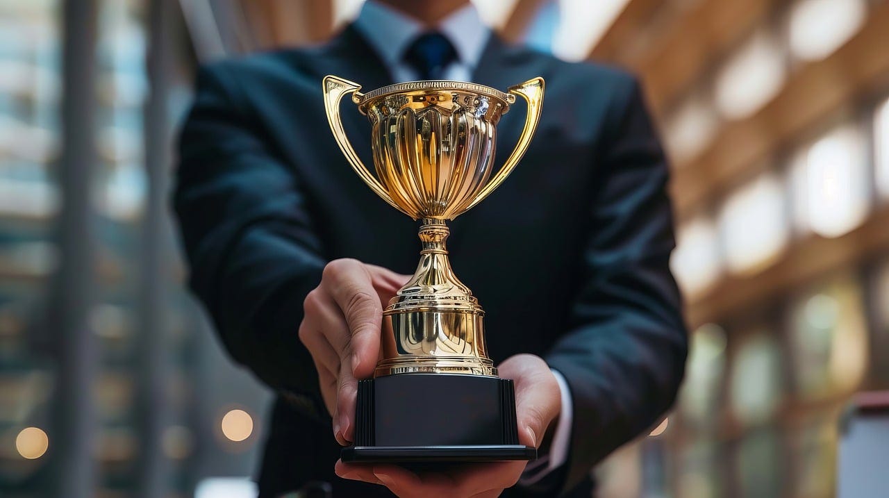 Man in suit holding a trophy in the foreground. He and the big-city background are blurred. The trophy is a gold chalice on a pedestal.