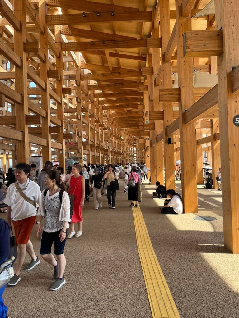 A walkway under a slightly curved wooden ceiling a few stories high, supported by wooden pillars and beams. There are dozens of people walking along the walkway, and a few people are sitting by pillars for shade.