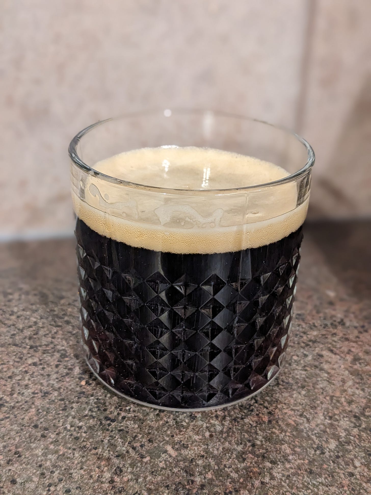 a glass of dark brown beer sits on a kitchen counter. the beer has light foam on top.