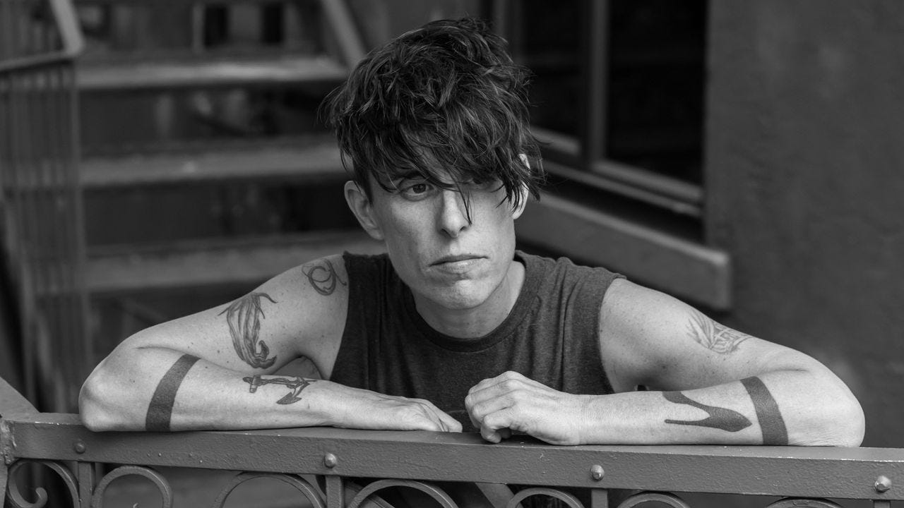 Black and white photo of a white person with messy dark hair over sincere eyes wearing a dark tank top, tattooed arms resting over a wrought iron fence