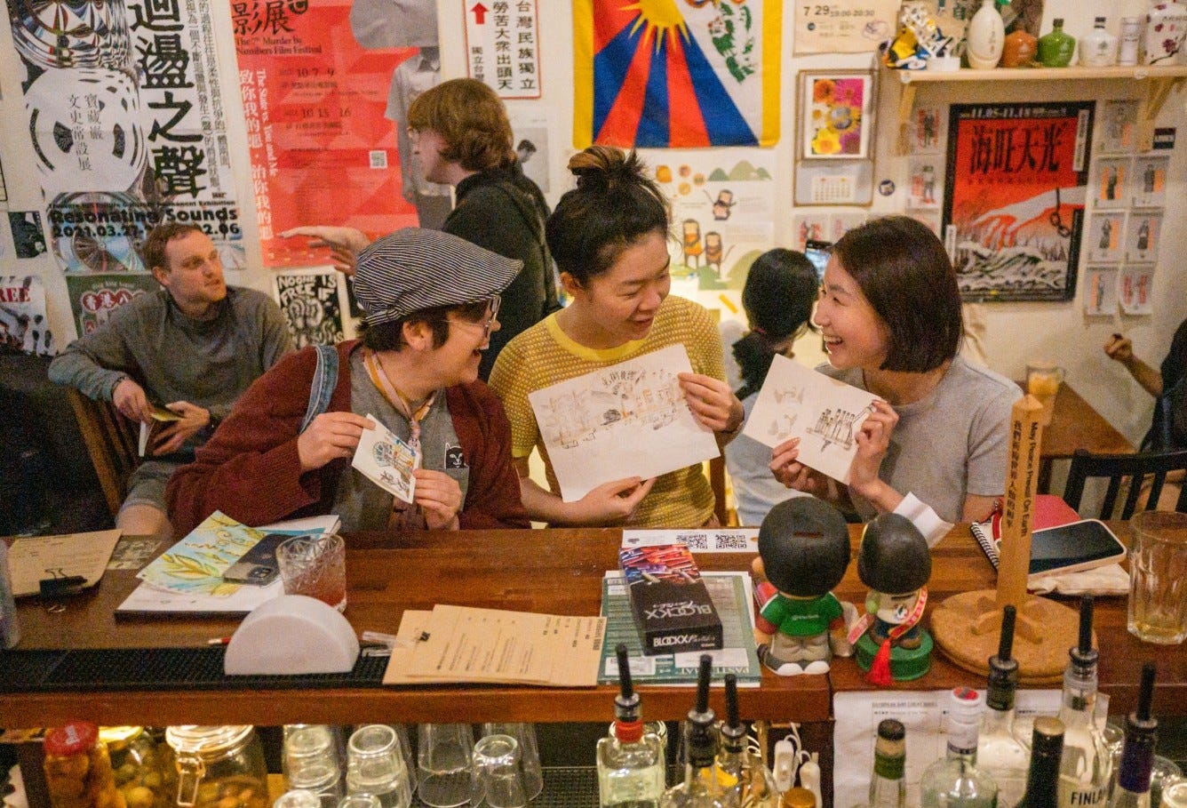 Three women laughing together seated at a bar, holding up their artwork. The view is from the barkeeper's side, with bottles visible. The walls of Daybreak are filled with social advocacy art. (Image contributed by Philip So)