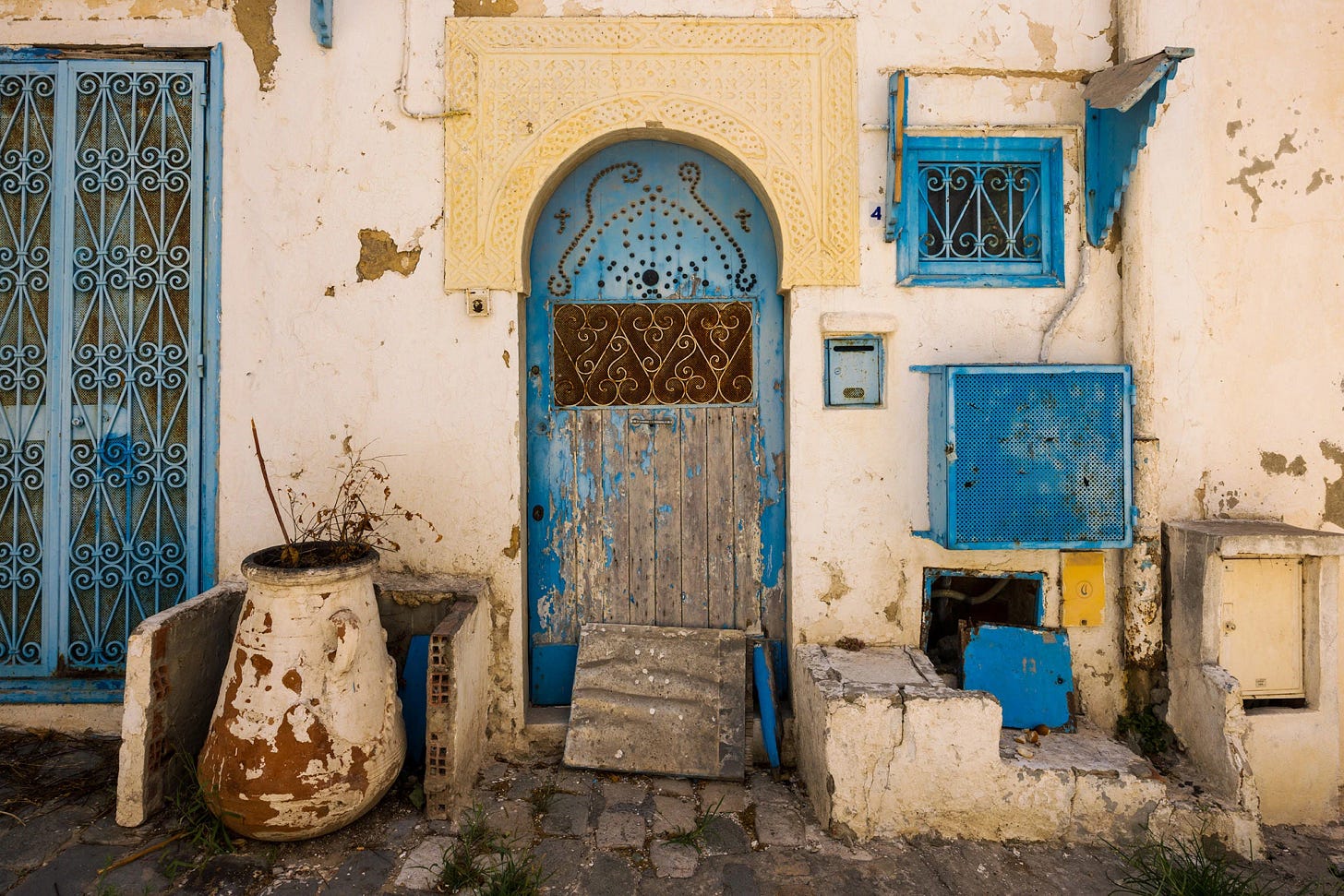 A weathered doorway in Sidi Bou Said, Tunisia, with peeling blue paint, carved plasterwork, and mismatched utilities framed by sun-faded walls. Signs of daily life—patched masonry, an old urn, and improvised repairs—reveal the lived-in texture that gives the village its enduring beauty and authenticity.