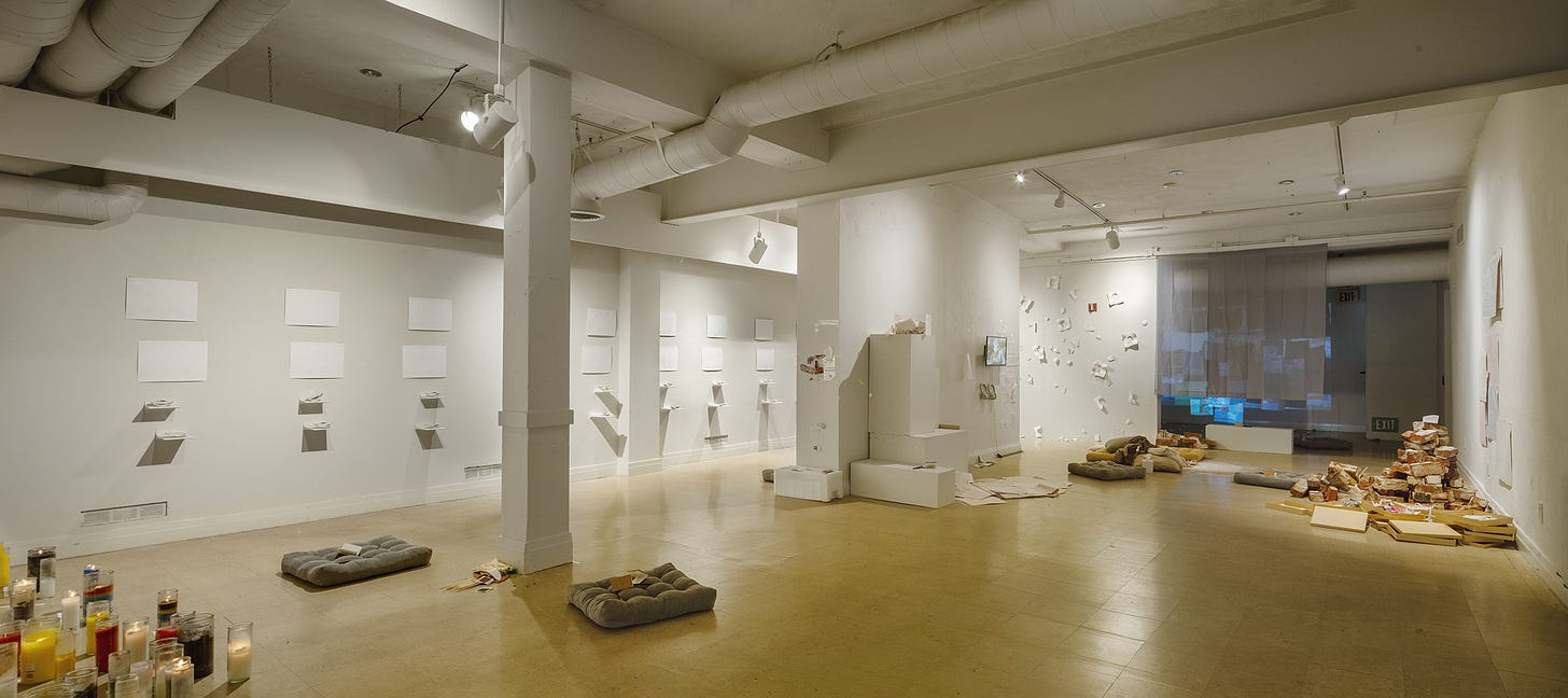 Wide view of a long white gallery. Small pale works are mounted in a grid along the left wall. In the middle distance, a round area rug with floor cushions and folded throws. Toward the back, a pile of reclaimed bricks and a low table. The floor is polished concrete.