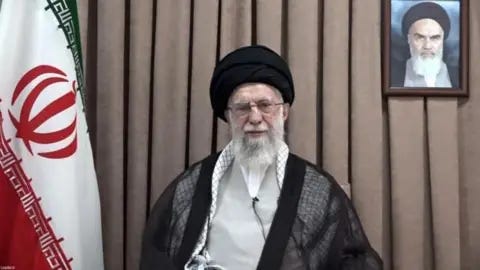 Reuters Ayatollah Ali Khamenei sits, wearing black and white, before a brown curtain with a large Iranian flag to his right and a portrait of Ayatollah Ruhollah Khomeini, Iran's founding father, to his left. 