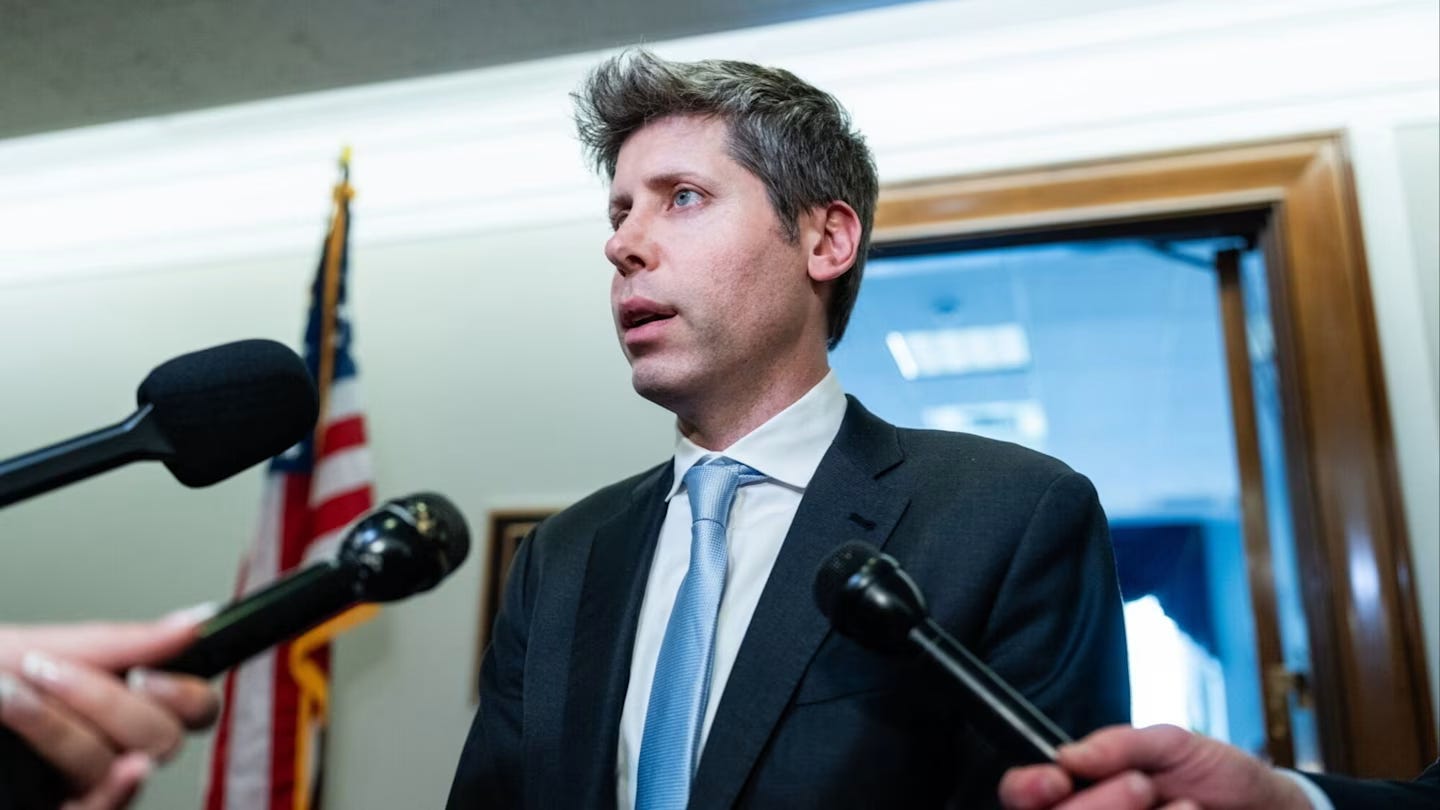 Sam Altman speaks to reporters in a hallway, with several microphones held toward him and a US flag in the background.